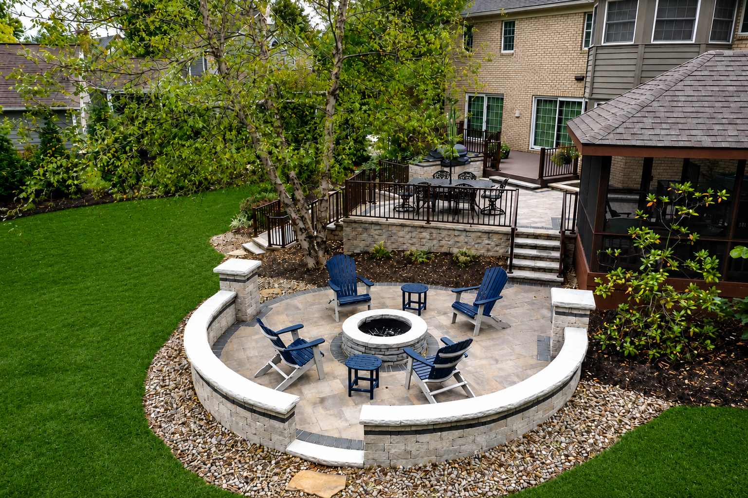 Stone patio with built-in stone firepit in foreground. Behind, up some stairs, a raised stone patio with a built-in grill and patio furniture that leads to residential home. Green lawn surrounds the patio tiers.