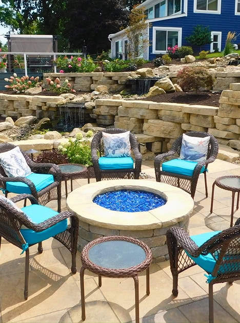 Stone firepit surrounded by lawn chairs. A stone retaining wall with waterfall in the background.
