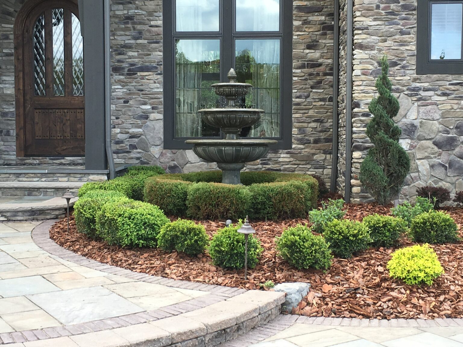 Landscape design and build for a front entry with tiered water fountain, shaped shrubs, mulch beds, paver walkway, and stone home exterior in Hudson, Ohio.